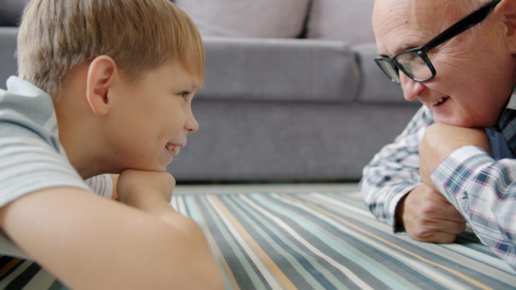 Grandfather and grandson smiling at each other, lying on the floor at home.